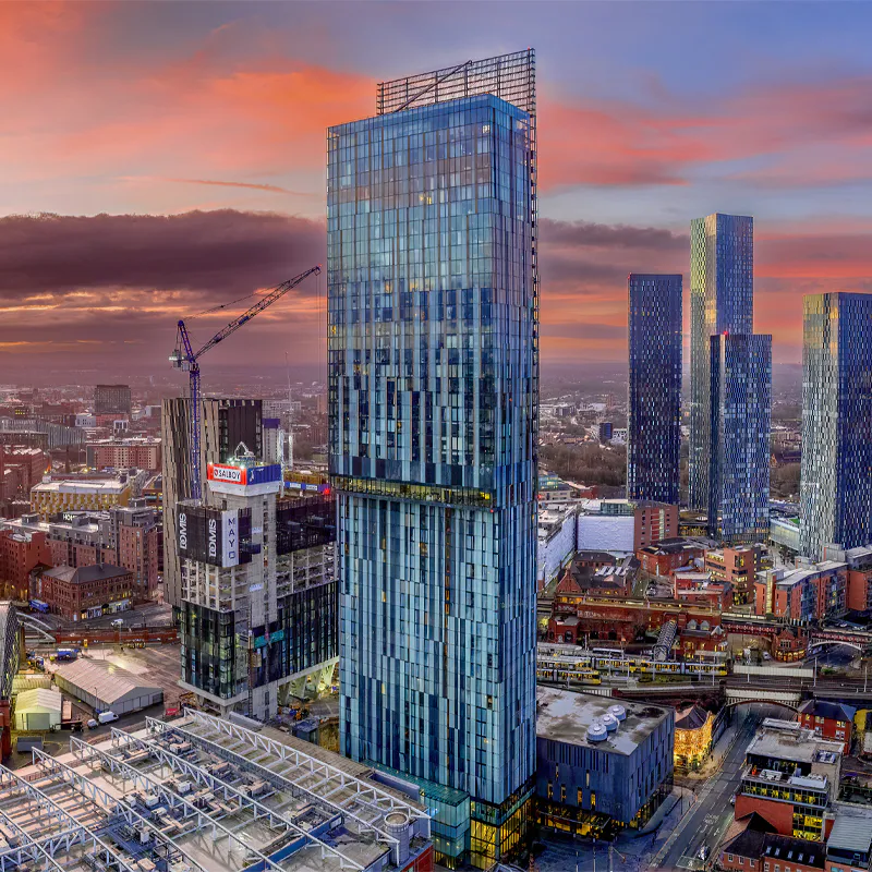 Manchester cityscape with high-rise buildings and construction cranes under a dramatic sunset sky.
