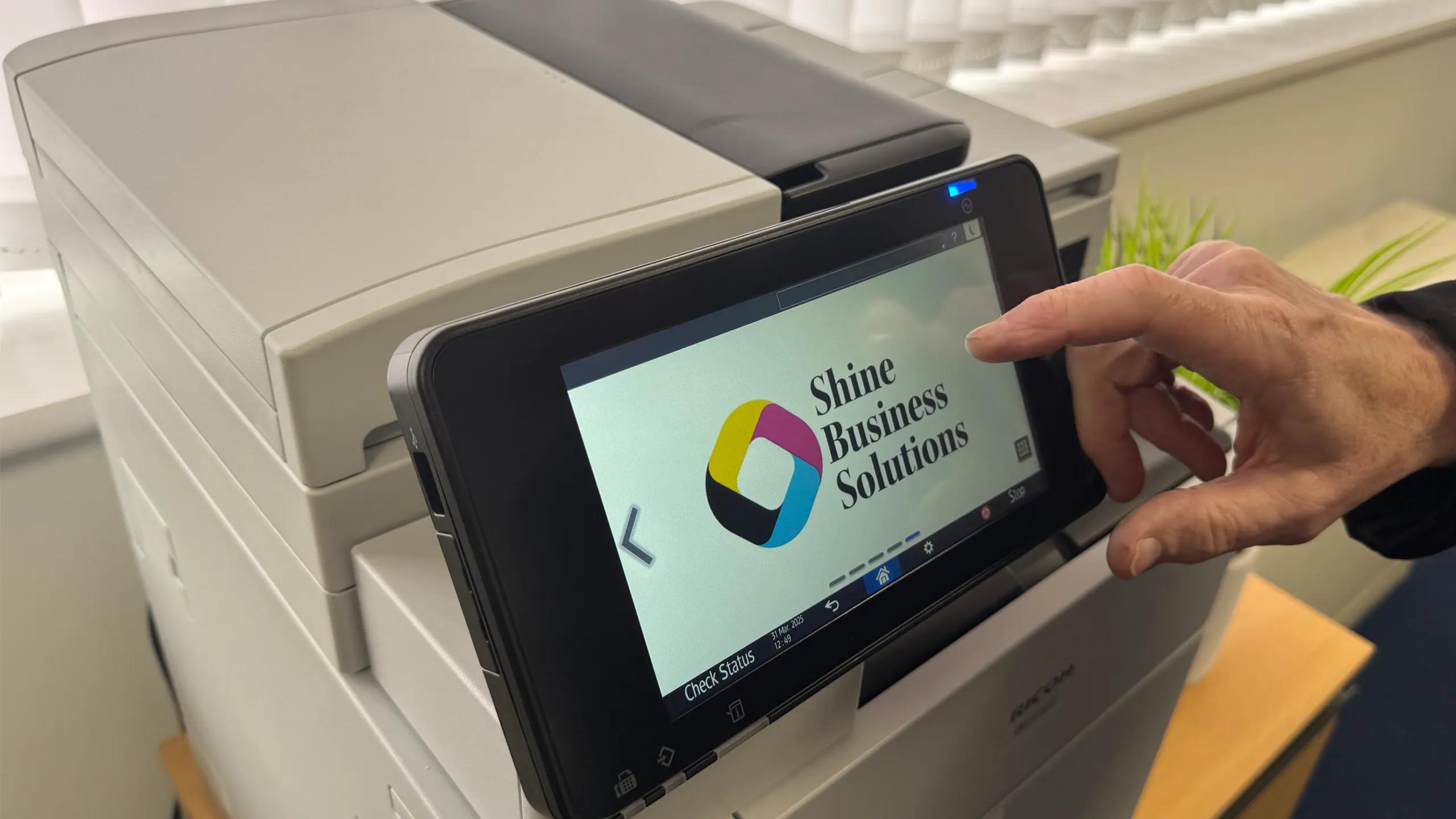 Hand on a printer touchscreen displaying the Shine Business Solutions logo, highlighting managed print services (MPS).